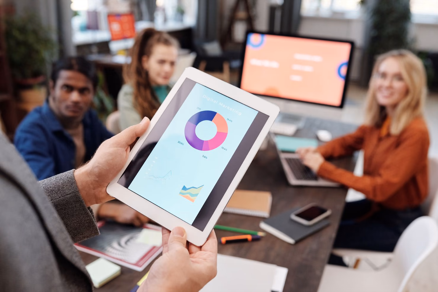 A team at a meeting table while one person holds a tablet showing a digital marketing donut chart.
