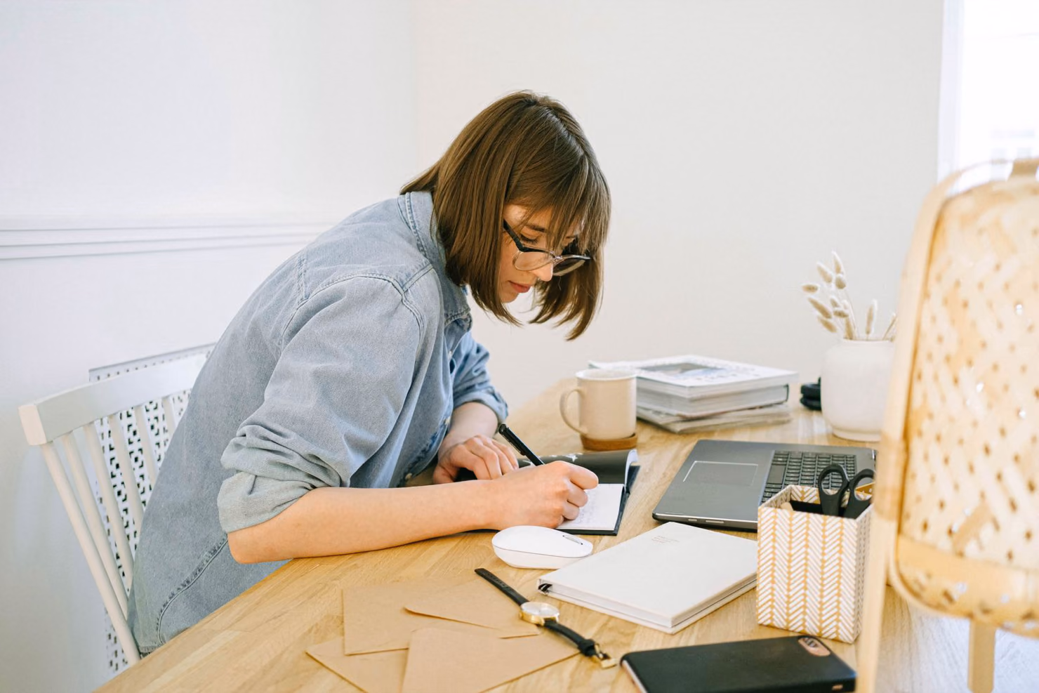 A woman at a wooden desk writing notes in a notebook beside an open laptop, planning content.
