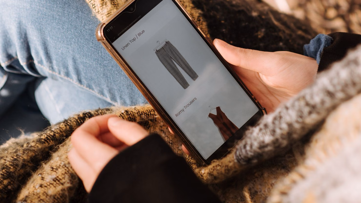 A shopper browsing a clothing product listing on a smartphone while sitting in a wicker chair with a blanket.