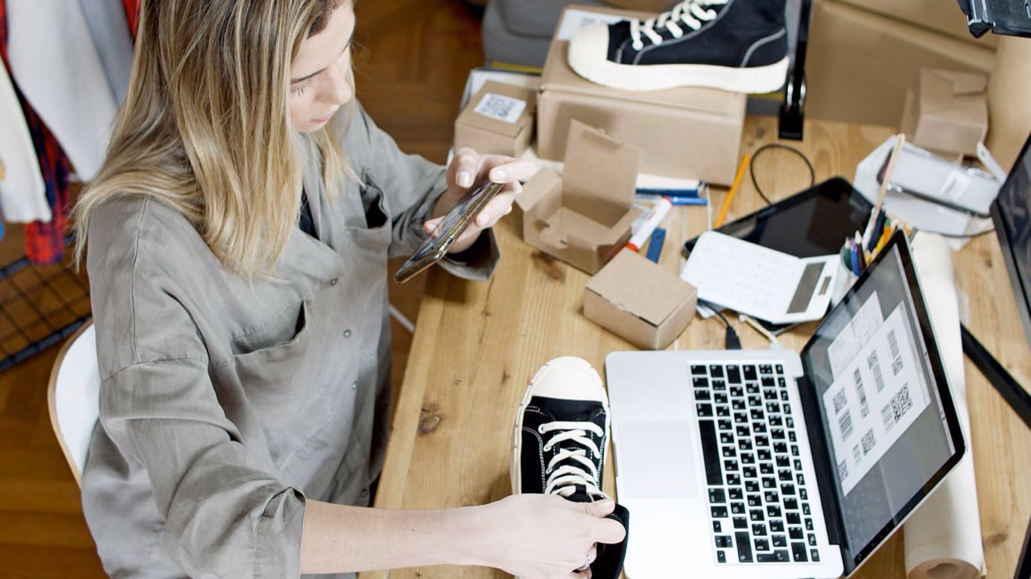 A small online seller photographing a black high-top sneaker with a phone at a desk covered in shipping boxes and a laptop showing product labels.