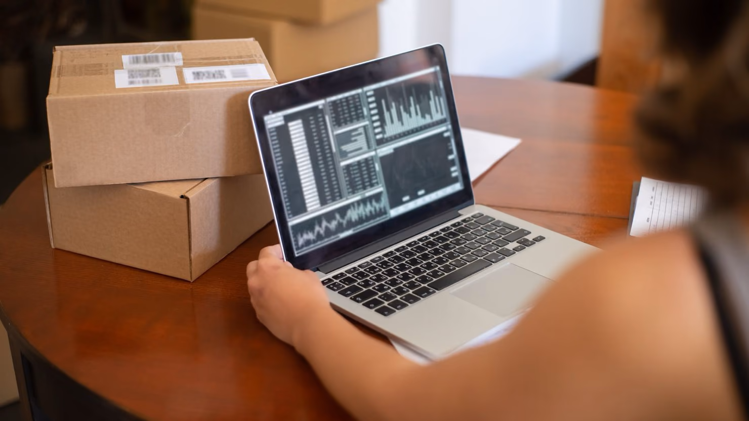 An online store owner reviewing sales charts on a laptop beside a stack of packed shipping boxes on a wooden table.