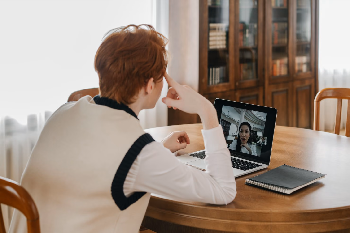A person at a wooden table on a video call with a developer shown on a laptop screen, with a notebook beside the laptop.