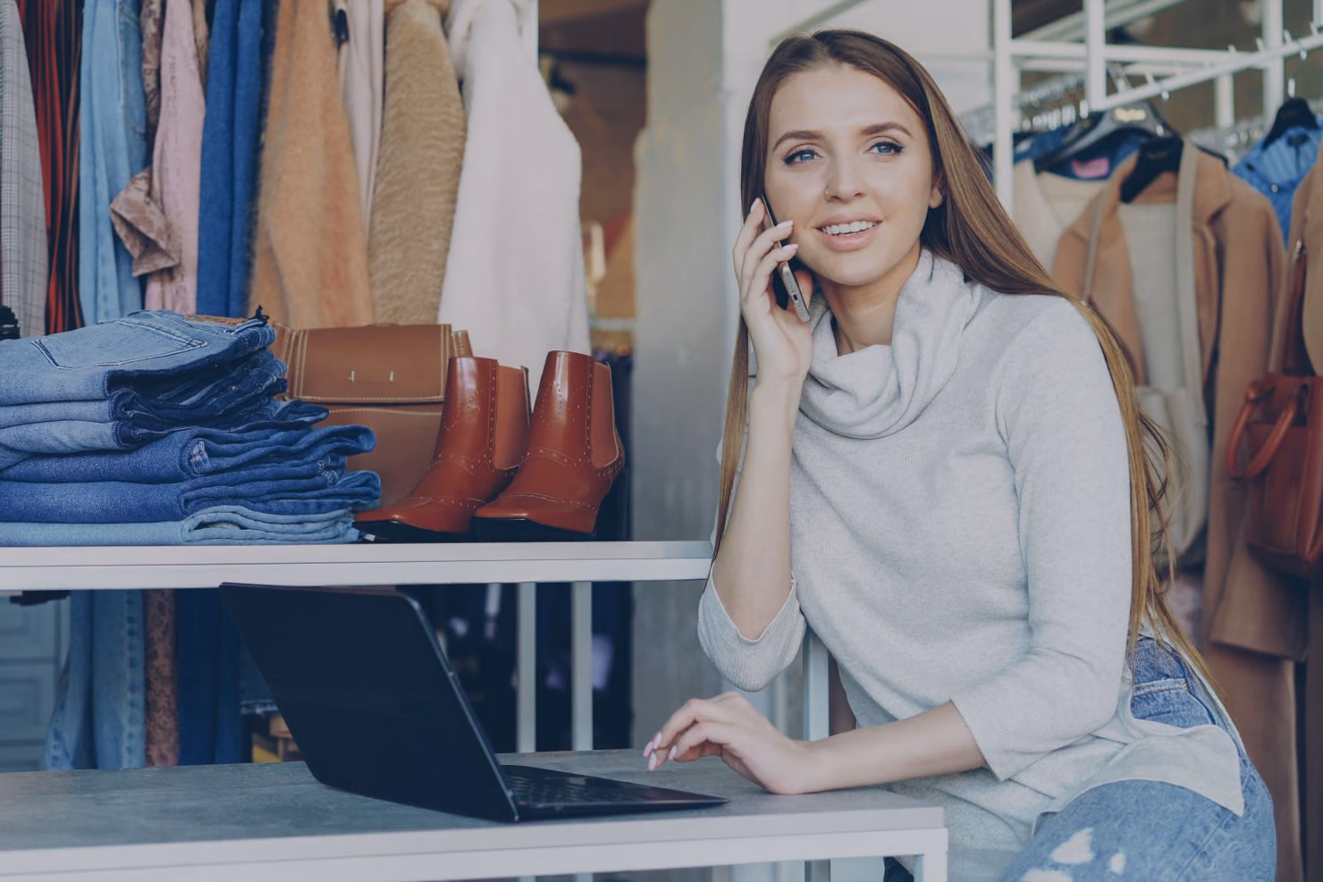 A clothing shop owner on the phone beside a laptop, with folded jeans, boots and a bag on the shelf next to her.