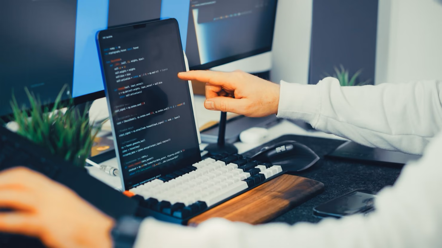 A developer pointing at lines of code on a tablet screen propped above a mechanical keyboard, with two monitors and a plant in the background.