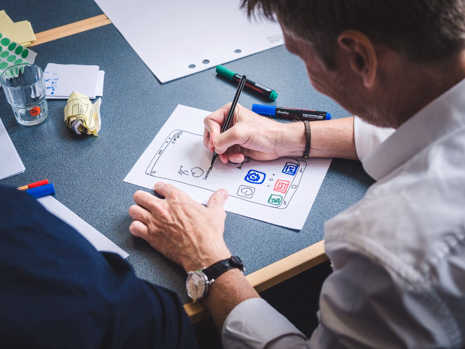 A man sketching the screens of a mobile app on paper with felt pens at a workshop table, with sticky notes and a glass of water beside him.
