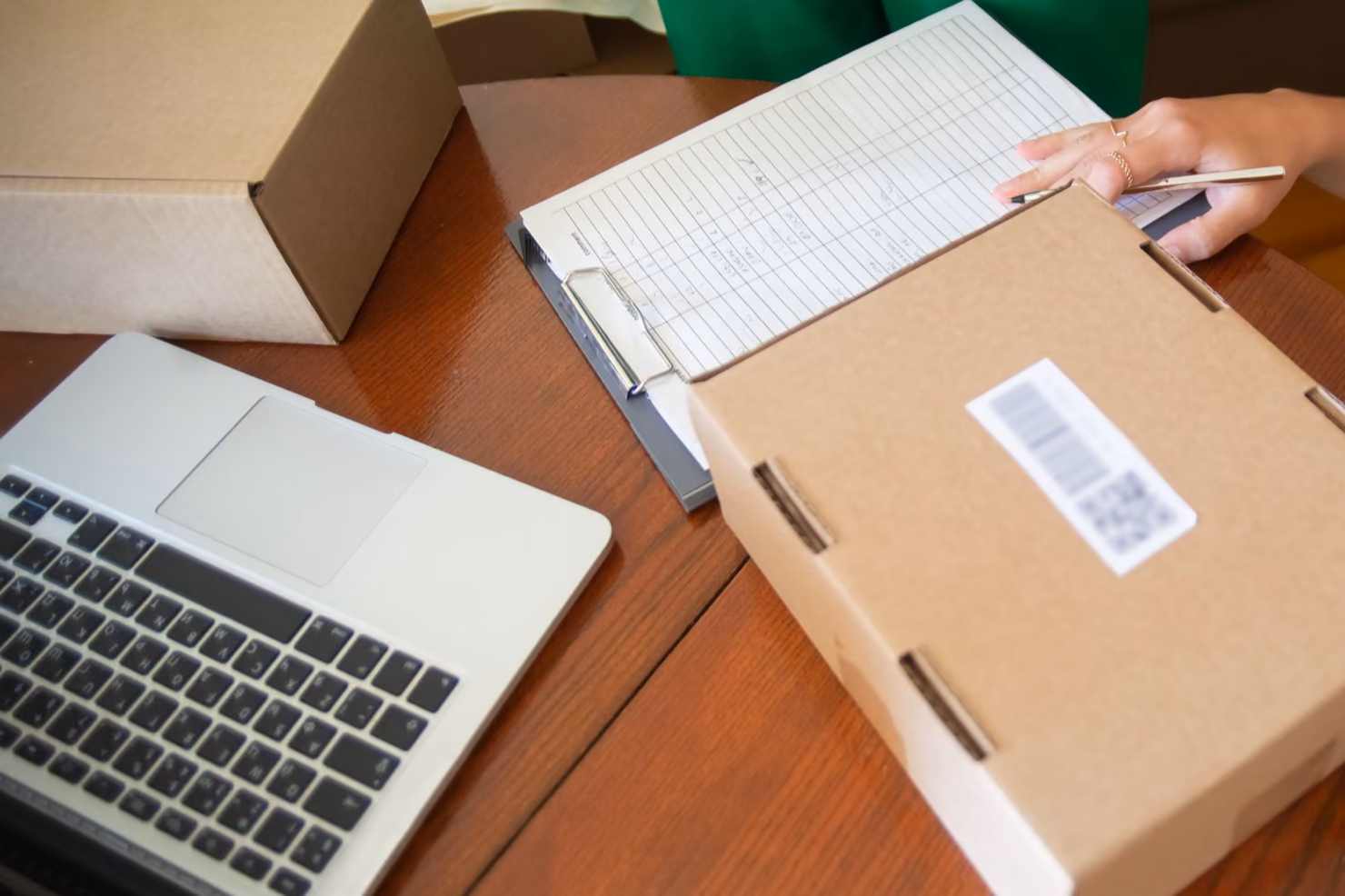 Overhead view of a small business packing table with a labelled shipping box, an order list on a clipboard, a laptop and packing tape.
